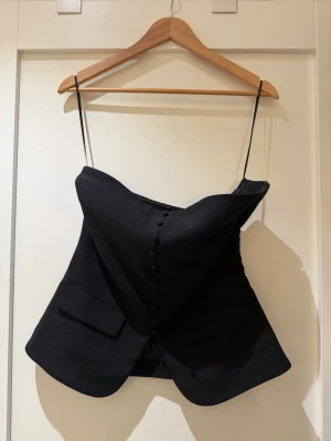 Svart korsettopp med knappar GinaTricot - Trendig svart korsettopp med smala axelband och dekorativa knappar framtill. Toppen har en unik, skräddad form med fickdetaljer och lätt utsvängd nederkant. Perfekt för dig som vill sticka ut med en edgy och stilren look.