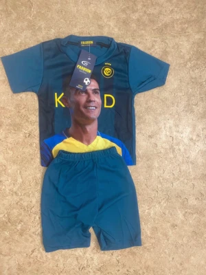 Blått fotbollsställ med tryck - Säljer ett blått fotbollsställ från Fashion med kortärmad tröja och matchande shorts. Tröjan har tryck med spelarmotiv, gula detaljer och klubbmärke på bröstet. Perfekt för dig som gillar fotboll och vill sticka ut på planen.