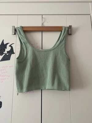 Ljusgrön ribbad linnetopp - En ljusgrön croppad linnetopp med breda axelband och rundad halsringning. Toppen är ribbad och har en enkel, stilren design som passar perfekt till jeans eller kjol. Materialet känns mjukt och stretchigt för en bekväm passform.