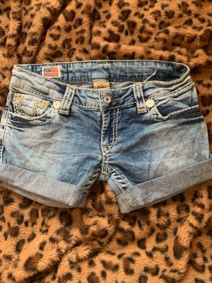 Blå jeansshorts från True Religion - Snygga blå jeansshorts från True Religion med uppvikta benslut och kontrastsömmar. Shortsen har klassiska fickor fram och bak, samt dekorativa broderier på bakfickorna. Perfekta för sommaren och ger en avslappnad streetstyle-look.
