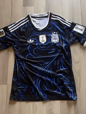 Argentina Adidas fotbollströja svart/blå (S) - Snygg svart fotbollströja från Adidas med blått mönster och vita detaljer. Tröjan har FIFA- och AFA-loggor på bröstet, samt tre vita ränder på axlarna. Tillverkad i ett lätt och snabbtorkande material, perfekt för träning eller match.