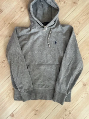 Grå Ralph Lauren Hoodie - Klassisk grå hoodie från Polo Ralph Lauren med mörkblå logga broderad på bröstet, Inga fläckar och hål med fåtal skador som inte syns på bilden. Hoodien har huva med vita dragsnören och en stor magficka. Tillverkad i mjukt bomullsmaterial och har ribbade muddar vid ärmslut och nederkant. Kontakta mig vid eventuella frågor. Pris kan förhandlas vid snabb och smidig affär 😊💫