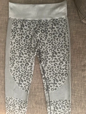 Grå leopardmönstrade träningstights - Snygga träningstights i grått med leopardmönster och bred ribbad midja. Tightsen har ribbade detaljer på sidor och benslut samt scrunch-effekt bak. Tillverkade i stretchigt material som passar perfekt för gym eller yoga.