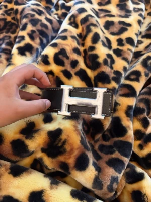 Bälte med H - Snyggt svart skärp med det ikoniska H-spännet i silverfärgad metall. Bältet har tydliga vita sömmar längs kanterna och är gjort i skinn. Perfekt accessoar för att lyfta vilken outfit som helst. Den är skrapad
