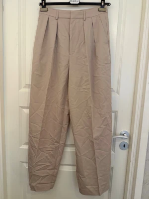 Beige vida kostymbyxor med pressveck - Snygga beige kostymbyxor med hög midja och vida ben. Byxorna har pressveck framtill, bälteshällor och diskreta fickor bak. Perfekta för en clean och stilren look. Materialet känns lätt och har en klassisk kostymkänsla. Oanvänd med prislapp. 