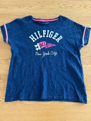 Blå Hilfiger t-shirt med tryck - Mörkblå t-shirt från Tommy Hilfiger med tryck i vitt och rosa framtill där det står 'Hilfiger All Star New York City'. T-shirten har korta ärmar med rosa och vita detaljer på ärmsluten och en rund halsringning. Perfekt för en avslappnad och sportig stil. Det står att det är storlek XL, men den sitter som en S eller mindre M.