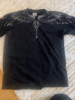 Svart t-shirt med vingmönster från Marcelo Burlon - Säljer en svart t-shirt från Marcelo Burlon med ikoniskt vingmönster i vitt och grått runt halsen, både fram och bak. T-shirten har normal passform och är tillverkad i mjuk bomull. Perfekt för dig som vill sticka ut med en unik design.