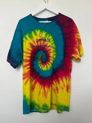 Färgglad Tie-dye T-shirt - En färgglad tie-dye t-shirt med spiral i blått, rött, gult och grönt. Tryck framtill med texten 'Jamaican Me Crazy' och en liten illustration av en buss, palmer och sol. Större och mysig tröja. Använd men i mycket fint skick. Storlek XL. 