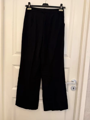 Svarta vida kostymbyxor - Säljer ett par svarta kostymbyxor med hög midja och vida ben. Byxorna har pressveck framtill, bälteshällor och sidfickor. Perfekta för en clean och stilren look. Materialet ser ut att vara en mjuk och följsam mix, troligtvis polyester eller viskos.