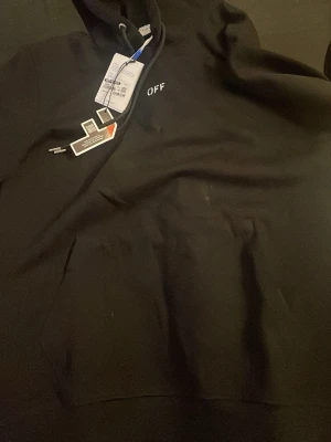 Svart hoodie från Off-White - Säljer en svart hoodie från Off-White med minimalistisk vit 'OFF'-logga på bröstet. Klassisk huva med dragsko och stor magficka framtill. Hoodien har en clean och stilren look, perfekt för dig som gillar streetwear och exklusiva märken.