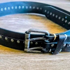 Svart bälte med plattar nitar och dubbla spännen - Snyggt svart bälte med dubbla metallspännen och silverfärgade nitar längs hela remmen. Bältet har en cool och edgy look som passar perfekt till jeans eller kjol. Tillverkat i skinn för en slitstark känsla.