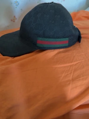 Svart keps från Gucci med GG-mönster - Snygg svart keps från Gucci med klassiskt GG-mönster över hela kepsen. På sidan finns en grön och röd rand som ger den en ikonisk touch. Justerbar baktill för perfekt passform. Tillverkad i bomullsmix för en bekväm känsla.