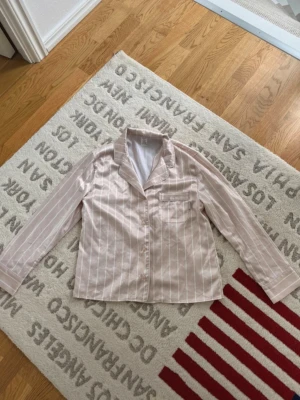 Randig pyjamasskjorta i ljusrosa - Snygg pyjamasskjorta i ljusrosa med vita vertikala ränder. Skjortan har klassisk krage, knäppning framtill och en bröstficka. Materialet känns mjukt och silkeslent, perfekt för mysiga kvällar hemma.