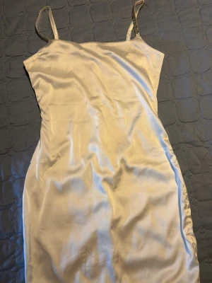 Vit satin klänning med strassband - Säljer en elegant vit långklänning i satin med tunna axelband dekorerade med strass. Klänningen har en slät, figurnära passform och är perfekt för dig som vill ha en stilren och glansig look. Perfekt till sommarens alla tillfällen!