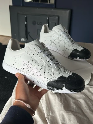 Vita sneakers med svart färgstänk - Unika vita sneakers med svart färgstänk och svartmålade tår. Skorna har lågt skaft, vita snören och en klassisk siluett. Materialet är slätt läder eller konstläder. Perfekta för dig som vill sticka ut med en kreativ och modern look.