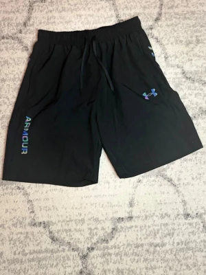Svarta träningsshorts från Under Armour - Säljer ett par svarta träningsshorts från Under Armour med elastisk midja och snörning. Shortsen har logga och text i regnbågsskimrande färg på ena benet. Perfekta för gymmet eller löprundan.