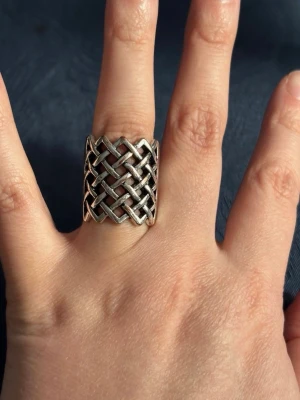 Bred Silverfärgad Ring med Flätmönster - Unik och bred ring i silverfärgad metall med ett detaljerat flätmönster framtill. Ringen har en robust känsla och sticker verkligen ut på fingret. Perfekt för dig som gillar statement-smycken och vill ha något som syns. I fint använt skick.
