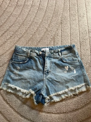 Blå lågmidjade jeansshorts med tryck och fransar - Säljer ett par blå lågmidjade jeansshorts från SR med råa fransade kanter och vitt tryck med hjärtan, revolver och texten 'cute but psycho' på ena benet. Shortsen har fem fickor och knappgylf. Perfekta för en avslappnad och cool stil. Kan klippas kortare❤️