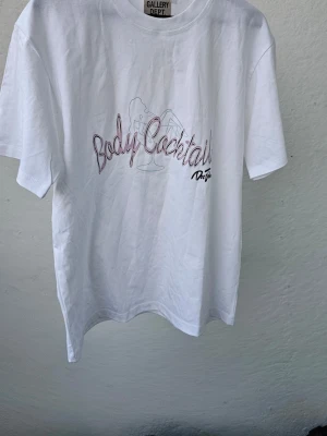 Vit t-shirt från Gallery Dept - Vit t-shirt från Gallery Dept med tryck 'Body Cocktail' på bröstet. Klassisk rund hals och korta ärmar. Materialet är mjuk bomull och passformen är normal. Perfekt för dig som gillar streetwear och coola prints.