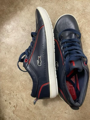 Mörkblå Lacoste sneakers med röd detalj - Snygga mörkblå sneakers från Lacoste med vita sulor och röda detaljer. Skorna har klassisk krokodillogga på sidan, mörkblå snören och är tillverkade i slätt läder. Perfekta för dig som vill ha en stilren och sportig look.
