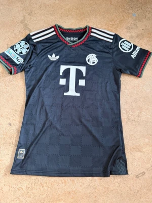 Svart Bayern München matchtröja Adidas - Snygg svart Bayern München matchtröja från Adidas med röda och vita detaljer. Tröjan har tryckta loggor, sponsortryck och Champions League-märke på ärmen. Namnet 'Olise' och nummer 17 på ryggen. Tillverkad i lätt och ventilerande material, perfekt för fotboll.