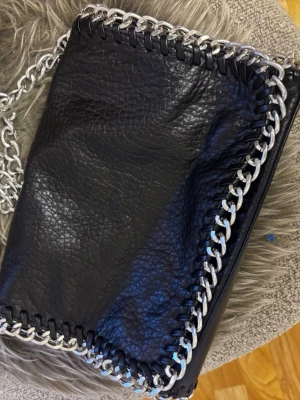 Svart handväska med kedjedetaljer Tiamo - Snygg svart handväska från Tiamo i fejkläder med grova silverfärgade kedjedetaljer runt kanterna och som axelrem. Köpt på Scorett!