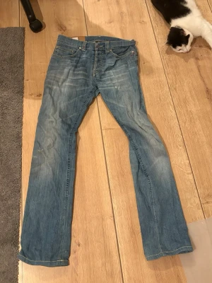 Blå raka jeans med klassisk tvätt - Säljer ett par blå dondup jeans med rak passform och klassisk tvätt. Jeansen har fem fickor, bälteshällor och knappgylf. Materialet är denim i bomull och de har subtila slitningar för en avslappnad look.