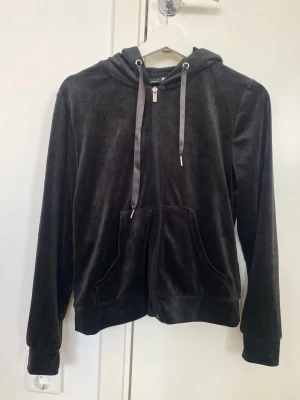 Svart velourhoodie med dragkedja - Mjuk och skön svart hoodie i velour med dragkedja framtill och stora fickor. Justerbar huva med breda snören ger en avslappnad vibe. Perfekt för dig som gillar en chill och stilren look. Passar till både jeans och mjukisbyxor.