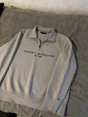 Grå halvzip sweatshirt Tommy Hilfiger - Snygg grå sweatshirt från Tommy Hilfiger med halvzip och broderad logga på bröstet. Tröjan har långa ärmar, ribbade muddar och en liten flagga på ärmen. Tillverkad i mjuk bomullsblandning och har klassisk collegekänsla. Storlek M. Väldigt bra skick