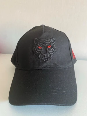 Svart keps med tigermotiv - Säljer en svart keps med broderad tiger på framsidan och röda detaljer i ögonen. Kepsen har böjd skärm och justerbar passform bak. Materialet är bomull och designen är stilren men ändå edgy.