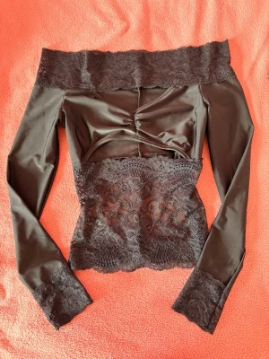 Svart offshoulder topp med spets - Trendig svart offshoulder topp med långa ärmar och snygga spetsdetaljer vid halsringning, midja och ärmslut. Toppen har en croppad passform och en lätt rynkad detalj framtill. Aldrig använd 