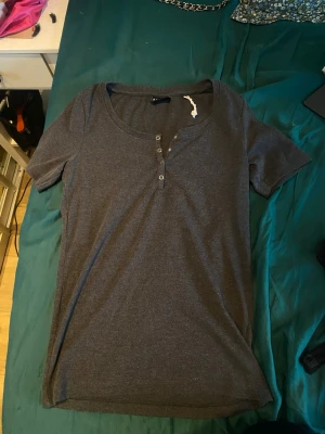 Mörkgrå t-shirt med knappar - En enkel mörkgrå t-shirt med korta ärmar och rund halsringning. Framtill finns en knappdetalj som ger en snygg touch. Perfekt basplagg till garderoben och lätt att matcha med olika stilar.