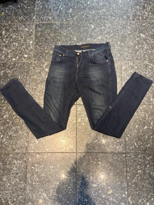 Mörkblå slim jeans från Nudie - Säljer ett par mörkblå slimjeans från Nudie Jeans med klassisk femficksmodell och guldfärgade sömmar. Jeansen har en smal passform och är tillverkade i stretchig denim för extra komfort. Perfekta för en avslappnad och stilren look. Dm för mer info