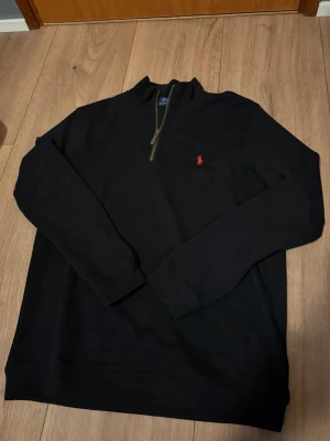 Svart Ralph Lauren quarter zip  - Svart långärmad tröja från Polo Ralph Lauren med quater-zip och klassisk röd broderad logga på bröstet. Tröjan har ribbade muddar och är gjord i mjukt bomullsmaterial. Perfekt för en clean och stilren look. Nyskick endast testad. Storlek M men passar även S