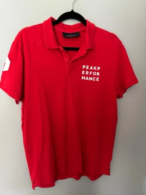 Röd Peak Performance pikétröja - Snygg röd pikétröja från Peak Performance med vit logga tryckt på bröstet. Klassisk krage och knappar framtill. Tillverkad i mjuk bomull som känns skön mot huden. Perfekt till jeans eller shorts för en avslappnad look.