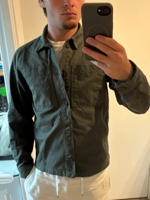 Olivgrön overshirt från Mouli - Säljer en stilren olivgrön overshirt från Mouli med två stora bröstfickor och knäppning framtill. Skjortan har klassisk krage och är tillverkad i ett slitstarkt bomullstyg. Perfekt att bära öppen eller stängd över en t-shirt. Mycket fint skick.