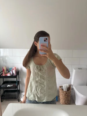 Ljusgrön blus från Odd Molly - Supersöt ljusgrön blus från Odd Molly med broderade detaljer och knytning vid halsen. Blusen har korta ärmar och är gjord i ett tunt, luftigt bomullstyg med hålmönster. Perfekt till jeans eller kjol för en fräsch look.