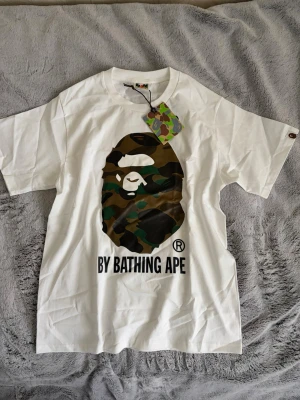 Vit BAPE t-shirt med camo-print - Säljer en vit t-shirt från BAPE med det klassiska apansikte-trycket i camouflage på bröstet och texten 'BY BATHING APE' under. T-shirten har rund hals och korta ärmar. Perfekt för dig som gillar streetwear och vill sticka ut.