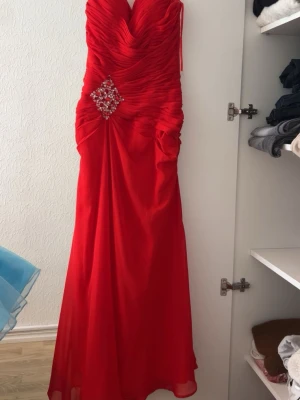 Röd långklänning med strassdetalj - Snygg röd långklänning med draperad överdel och v-ringning. Klänningen har en iögonfallande strassdetalj i midjan och är figurnära upptill med ett lätt utställd kjol. Perfekt för bal eller festliga tillfällen. Denna klänning formar höfterna extremt vackert.