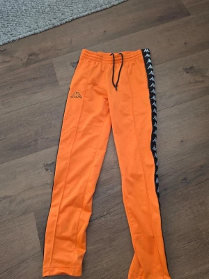 Orange mjukisbyxor från Kappa - Säljer ett par orange mjukisbyxor från Kappa med svarta och vita loggor längs sidorna. Byxorna har resår i midjan med svart dragsko och tryckknappar längs benen. Perfekta för en sportig och avslappnad stil. XS men passar som S.