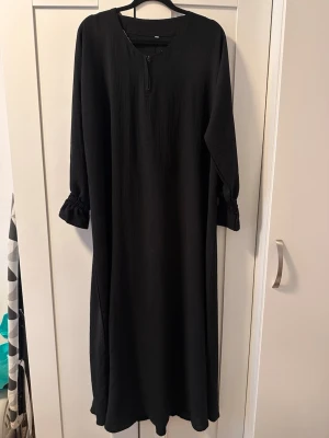 Svart långklänning med volangärm - En svart långklänning med rund halsringning och liten slits framtill. Klänningen har långa ärmar med volangdetalj vid ärmslutet och en lös, rak passform. Perfekt för dig som gillar enkel och stilren design.