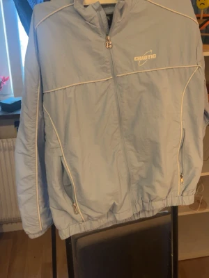  Chaotic tracksuit  - Ljusblå vindjacka från Chaotic med vita detaljer och dragkedjor. Jackan har hög krage, två fickor med dragkedja och vita kontrastsömmar som ger en sportig look. Materialet är lätt och passar perfekt för blåsiga dagar.