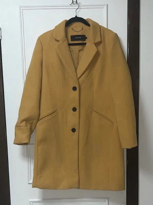 Senapsgul rock från Vero Moda - Stilren senapsgul rock från Vero Moda med tre svarta knappar framtill och klassisk krage. Rocken har två snedställda fickor och en rak passform som ger en snygg siluett. Perfekt för dig som vill sticka ut med färg i höst och vinter.