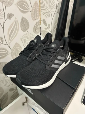 Svarta Adidas Ultraboost sneakers - Svarta Adidas Ultraboost sneakers med stickad ovandel, vita Boost-sulor och klassiska tre ränder i grått på sidan. Skorna har en sportig look med låg profil och bekväm passform. Perfekta för dig som gillar stilrena och funktionella sneakers.