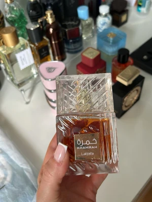 Lattafa Khamrah Eau de Parfum - Lattafa Khamrah är en lyxig parfym i en elegant, fyrkantig glasflaska med ett mönstrat lock. Flaskan har en varm bärnstensfärgad vätska och en guldfärgad etikett med både arabisk och engelsk text. Perfekt för dig som gillar exklusiva dofter och snygg design på parfymflaskan. Full förpackning, endast sprayad vid ett tillfälle. 