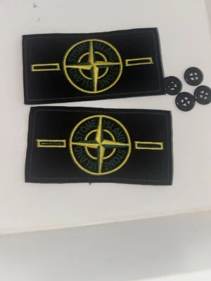 Stone Island badge bundle - Två svarta tygmärken från Stone Island med det klassiska gula och gröna kompasslogotypen broderad framtill. Märkena kommer med fyra svarta knappar för enkel fastsättning på plagg. Perfekt för att ge din jacka eller tröja en unik touch.
