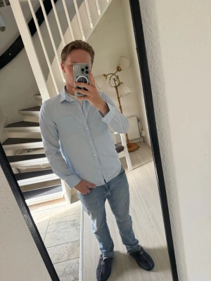 Ljusblå skjorta med knappar - Säljer en stilren ljusblå skjorta med klassisk krage och långa ärmar. Skjortan har knappar framtill och två bröstfickor. Perfekt till jeans eller chinos för en clean look.
