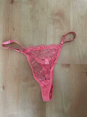 Rosa spetsstring med blommönster - Säljer en snygg rosa stringtrosa i spets med blommigt mönster och små rosetter framtill. Trosan har tunna, justerbara band i sidorna och är gjord i ett genomskinligt spetsmaterial som ger en feminin och trendig look.