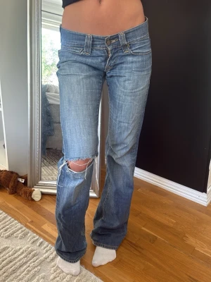 Blå slitna bootcut jeans - Säljer ett par blå bootcut jeans med slitning och hål på vänster knä. Jeansen har klassisk femficksdesign, låg midja och dragkedjegylf. Perfekta för en avslappnad och trendig look.