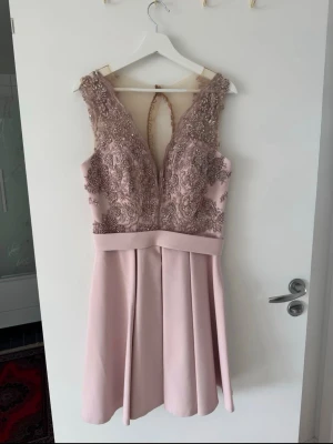 Ljusrosa spetsklänning med v-ringning - Supersnygg ljusrosa klänning med v-ringad spets upptill och markerad midja. Klänningen har vackra broderade detaljer, knappar i ryggen och en lätt utsvängd kjol. Perfekt för speciella tillfällen och ger en elegant look. Storlek är M men passar även S. 